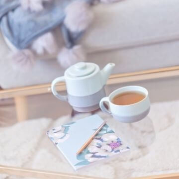 teapot, cup of tea, floral gratitude journal, sofa and comfy banket in the background
